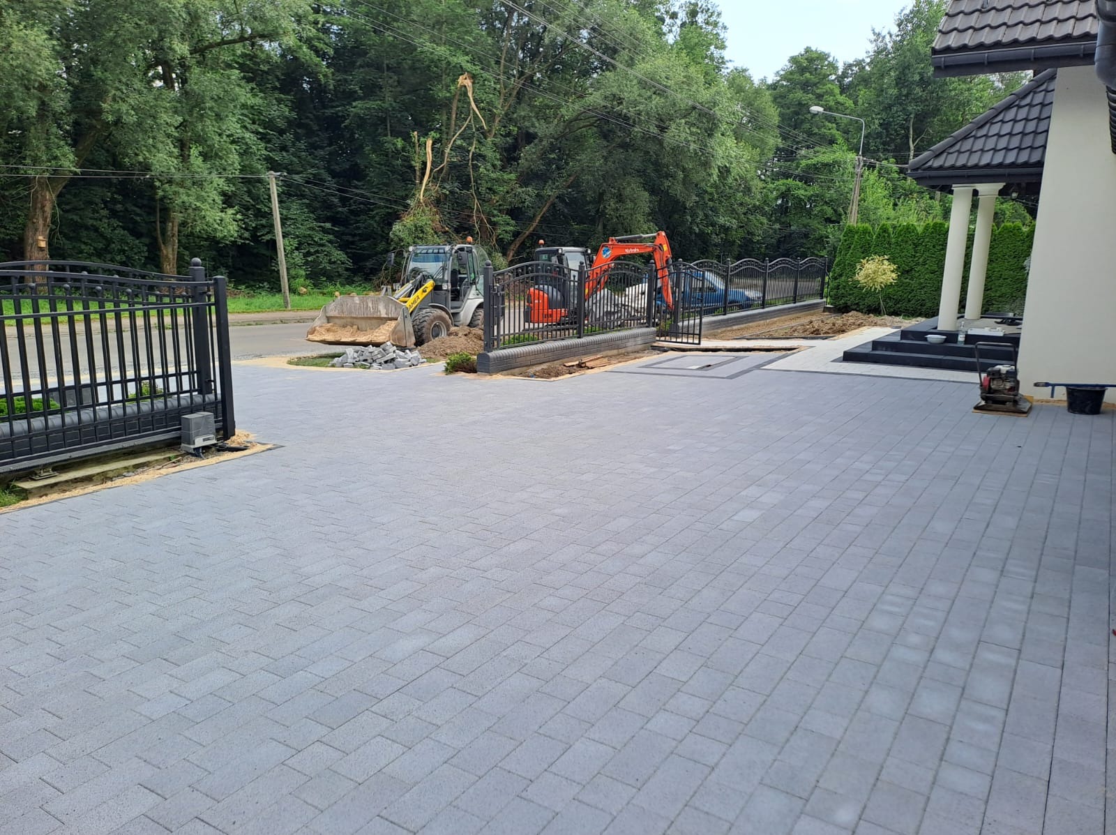 Ukończony podjazd z szarej kostki brukowej, widoczny wjazd z automatyczną bramą, w tle prace ziemne wykonywane koparkami i miniładowarką.