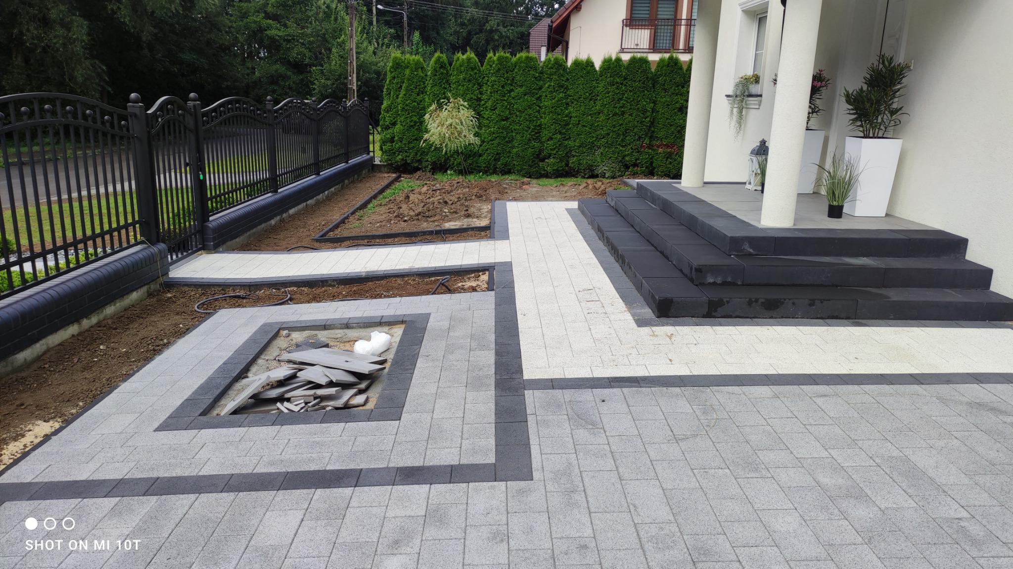 Ułożona szara kostka brukowa z ciemnymi obramowaniami prowadząca do schodów z czarnej kostki, widoczny fragment ogrodzenia i przygotowane miejsce pod nasadzenia.