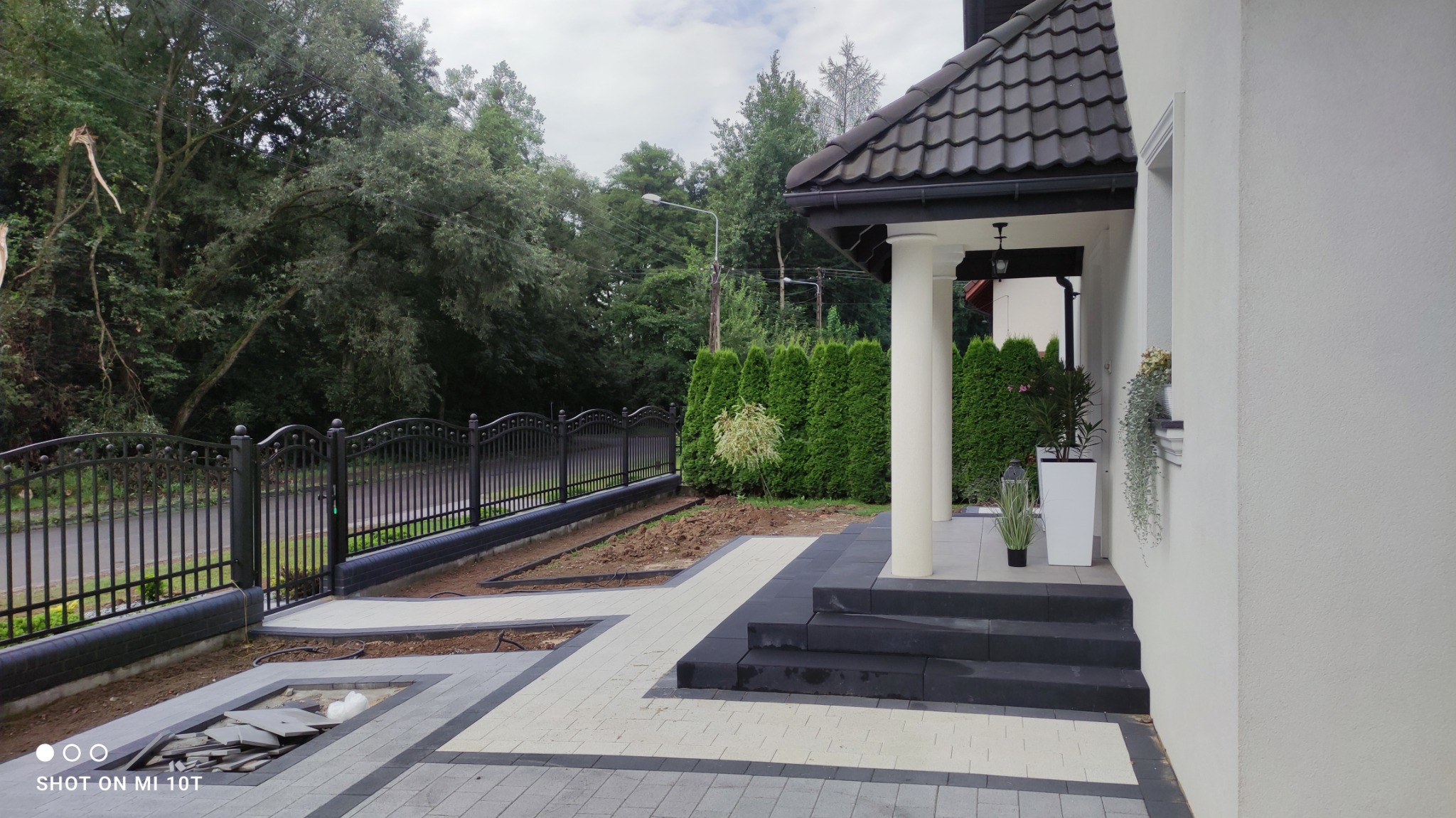 Nowoczesny podjazd z szarej kostki brukowej z czarnymi obramowaniami, prowadzący do schodów wejściowych i bramy z czarnym metalowym ogrodzeniem, na tle zieleni.