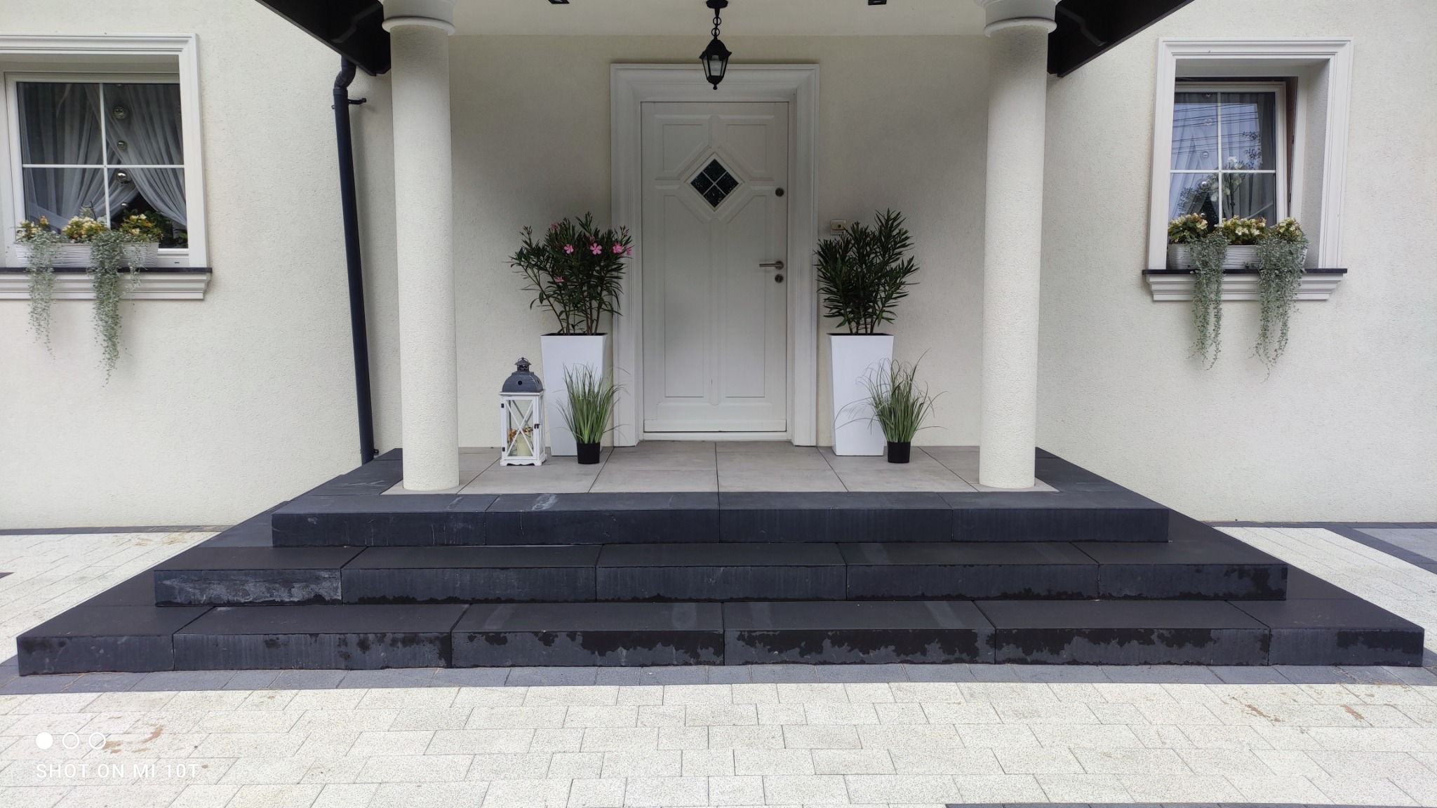 Eleganckie wejście do domu z czarnymi schodami z kamienia, jasną kostką brukową, kolumnami i białymi donicami z roślinami po bokach białych drzwi.