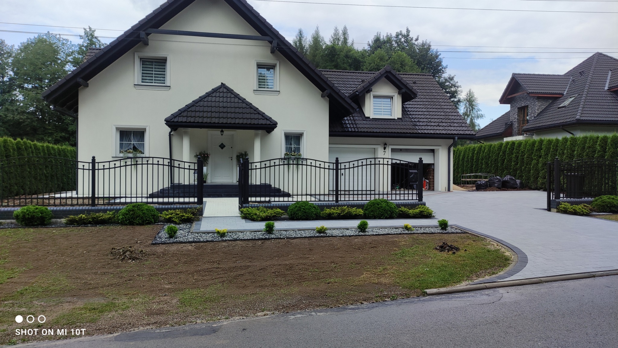 Nowoczesny dom jednorodzinny z kremową elewacją, czarnym dachem i czarnym, kutym ogrodzeniem, otoczony świeżo założonym trawnikiem i ozdobnymi krzewami, z wybrukowanym podjazdem.