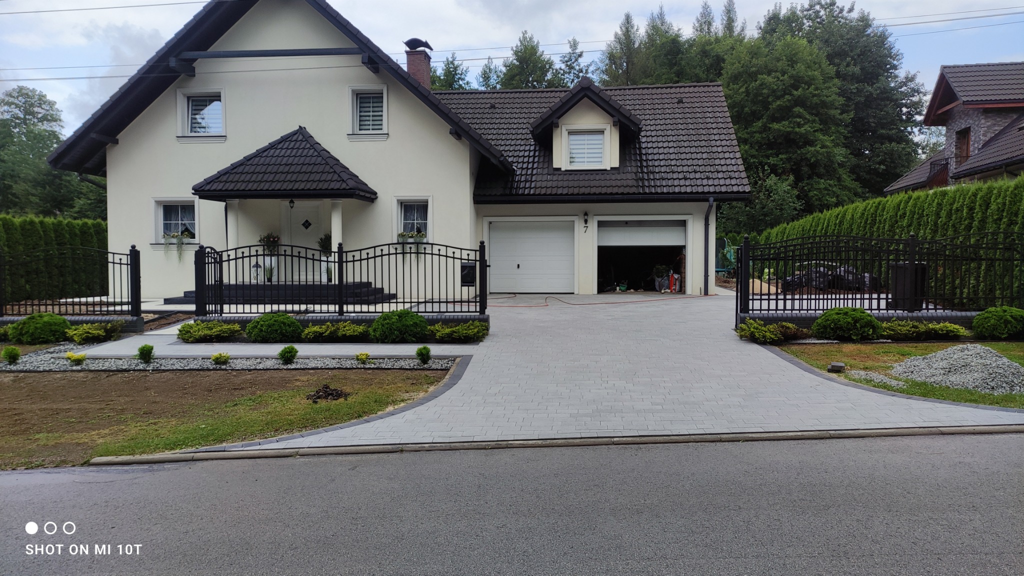 Nowoczesny podjazd z jasnoszarej kostki brukowej prowadzący do domu w Wiśle, otoczony czarnym ogrodzeniem i rabatami z niską zielenią, z otwartym garażem w tle.