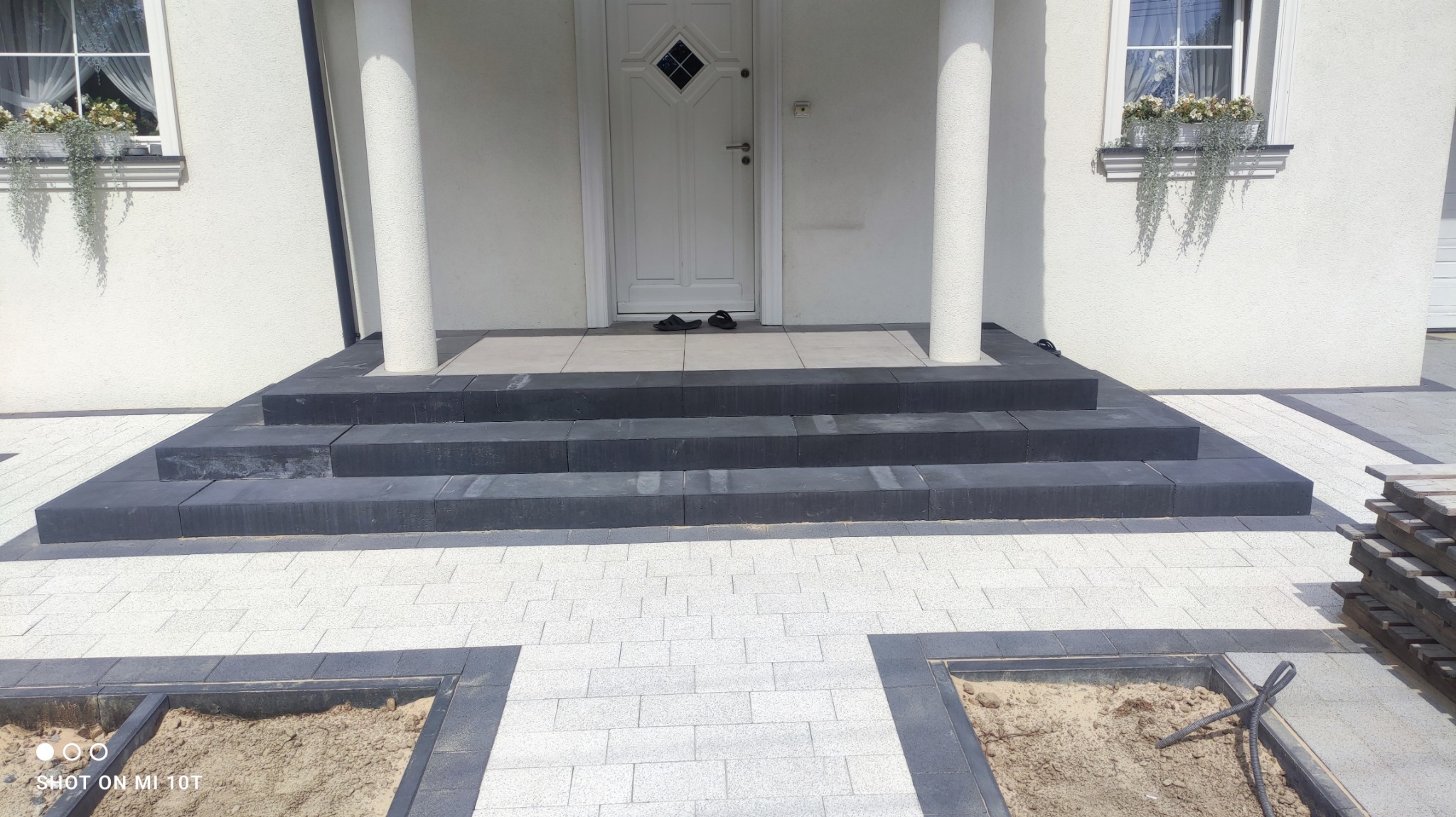 Nowe schody kamienne z ciemnej kostki prowadzące do wejścia domu, otoczone jasną kostką brukową w trakcie układania, widoczne puste przestrzenie przygotowane pod kostkę.