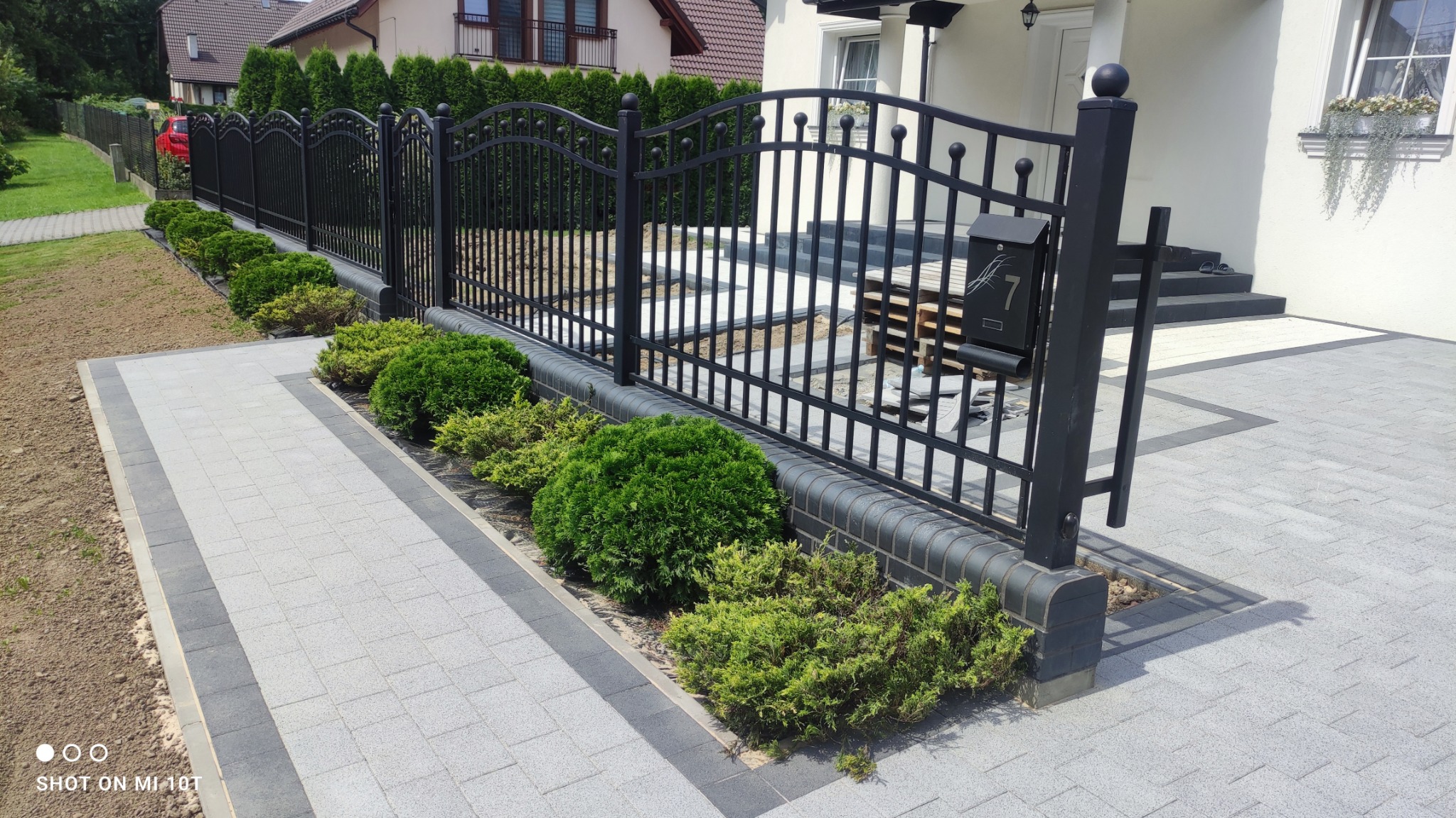 Nowoczesna posesja z szarą kostką brukową, czarnym metalowym ogrodzeniem z numerem 7 na skrzynce pocztowej i równo przyciętymi krzewami, widok z poziomu gruntu.