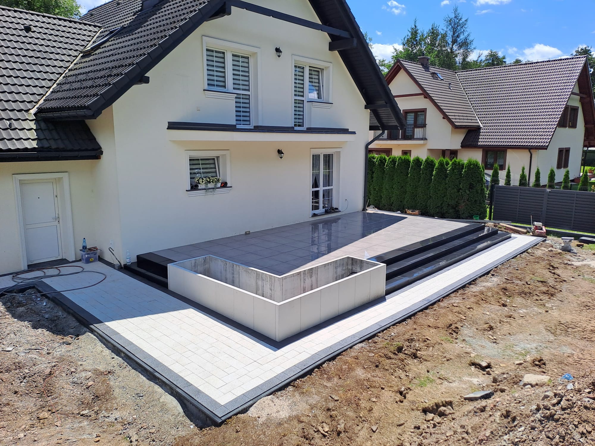 Nowoczesny taras z jasnoszarej kostki brukowej z ciemnoszarym obramowaniem, schody z czarnego kamienia prowadzące do drzwi balkonowych, biała donica betonowa, w tle biały dom z ciemnym dachem...