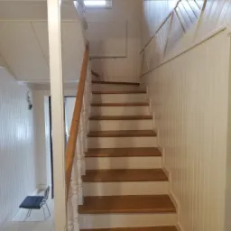 Drewniane schody z białymi balustradami i boazerią w jasnym wnętrzu z drewnianym sufitem, widoczne z dołu.