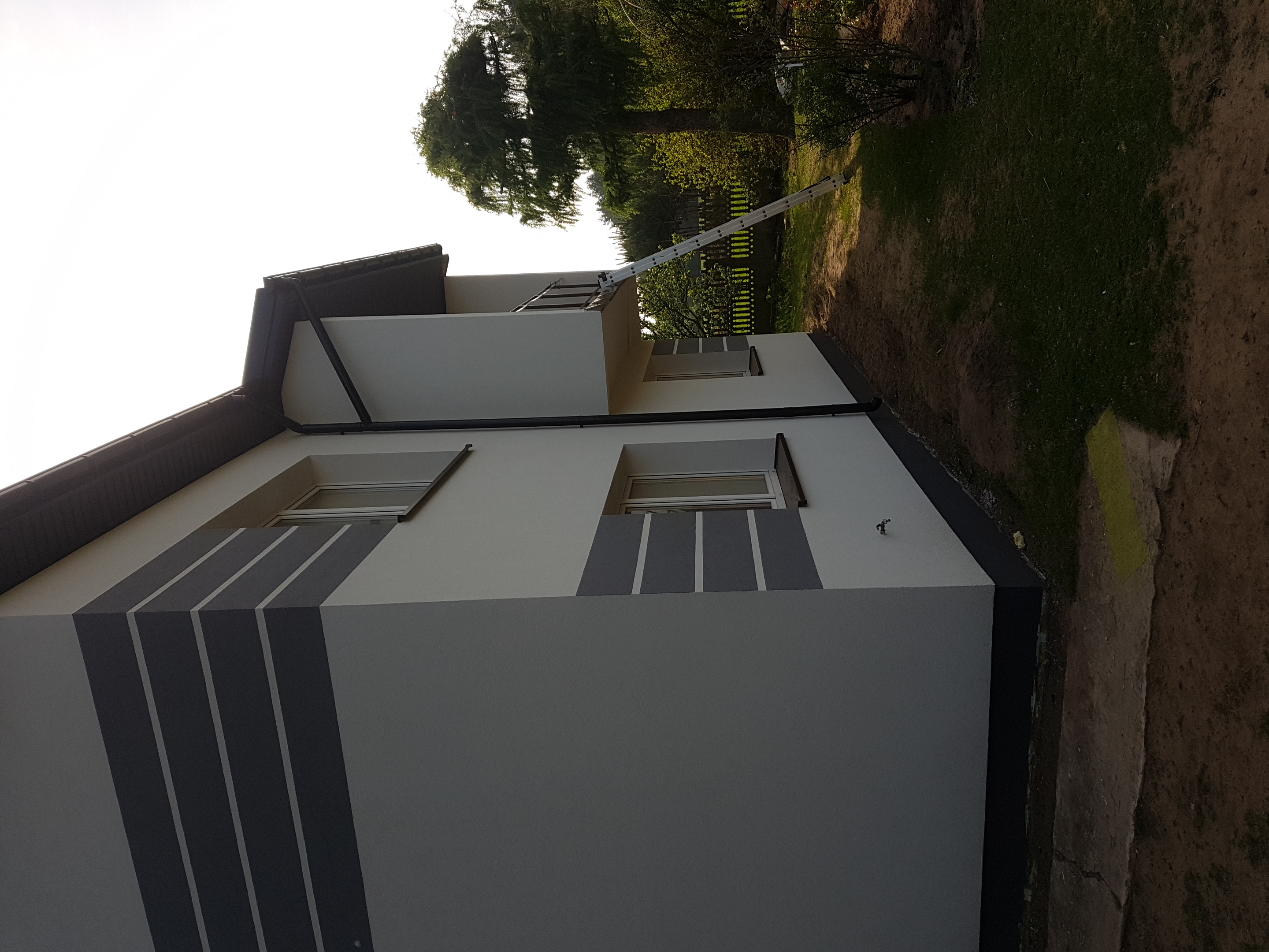 Nowoczesny dom jednorodzinny z jasnoszarą elewacją i ciemnoszarymi, pionowymi pasami dekoracyjnymi; widoczna aluminiowa drabina oparta o ścianę.