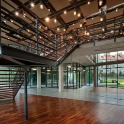 Wnętrze nowoczesnego budynku z drewnianą podłogą, betonowym sufitem podwieszanym z oświetleniem punktowym i stalowymi schodami z balustradą.