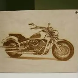 Wypalony laserowo wizerunek motocykla na jasnej, drewnianej płycie z widocznymi otworami montażowymi w rogach.