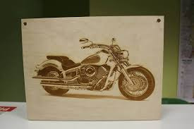 Wypalony laserowo wizerunek motocykla na jasnej, drewnianej płycie z widocznymi otworami montażowymi w rogach.
