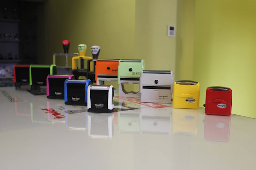 Różnokolorowe automatyczne pieczątki Trodat i Wagraf ustawione w rzędzie na białym blacie, z widocznymi odbiciami i zielonym tłem.