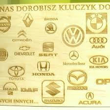 Tablica z wygrawerowanymi laserowo logotypami różnych marek samochodów na jasnym drewnie, prezentująca ofertę dorabiania kluczyków.