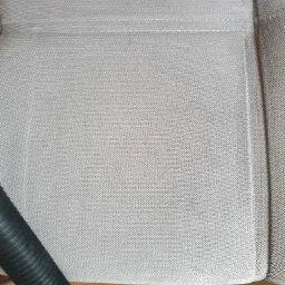 Fragment jasnej tapicerki kanapy z widocznymi śladami zabrudzeń przed czyszczeniem, z czarnym wężem odkurzacza w lewym dolnym rogu i napisem 'Po' w lewym górnym rogu.