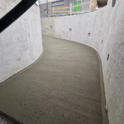 Świeżo wylana, gładka betonowa posadzka w zakrzywionym tunelu z surowego betonu, widoczne otwory technologiczne w ścianach, czarna rura instalacyjna pod sufitem, budynek w tle.