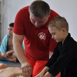 Instruktor w czerwonej koszulce z napisem 'Ratownik Medyczny' uczy chłopca w czarnym szlafroku wykonywania resuscytacji krążeniowo-oddechowej na manekinie podczas warsztatów dla dzieci.