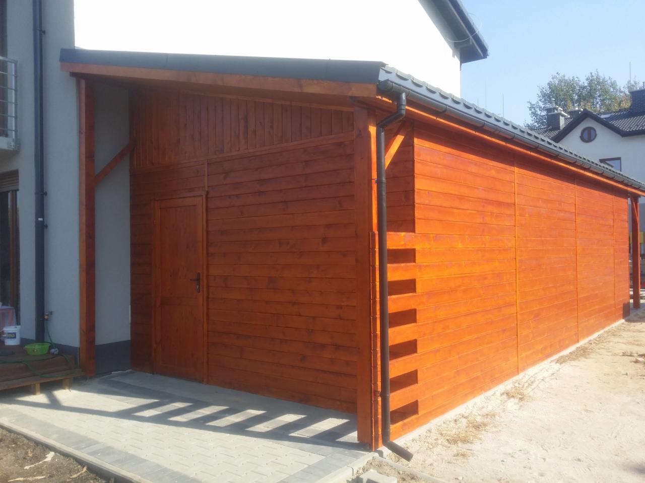 Nowoczesna, drewniana wiata garażowa z drzwiami, pomalowana na ciepły, brązowy kolor, z widocznym systemem rynnowym i kostką brukową przed wejściem.