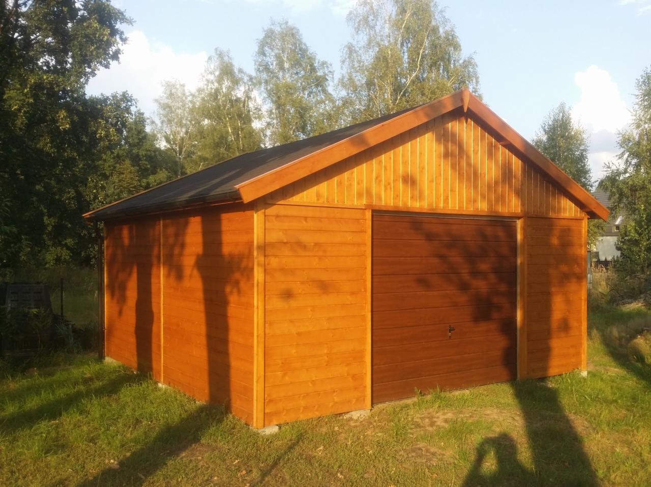 Drewniany garaż z brązową bramą segmentową, wykończony pionowo i poziomo ułożoną boazerią, na tle zieleni i drzew.