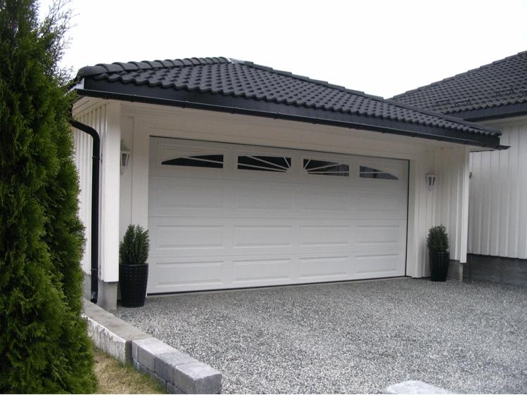Biała segmentowa brama garażowa z przeszkleniami w górnej części, czarny dach z dachówki ceramicznej, elewacja z białych desek, szary podjazd z drobnego kamienia.