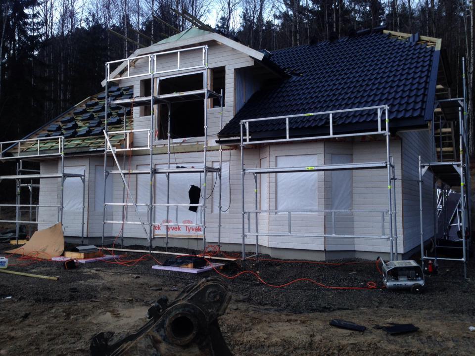 Dom w trakcie budowy z widocznymi rusztowaniami, częściowo pokrytym dachem i oknami zabezpieczonymi folią, otoczony leśnym krajobrazem.