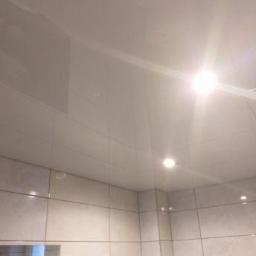 Sufity napinane sufit design - Błyszczący, biały sufit z widocznymi punktowymi lampami w łazience z jasnymi, kwadratowymi płytkami ceramicznymi na ścianach i lustrem.