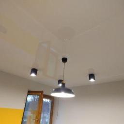 Sufity napinane sufit design - Błyszczący, biały sufit z zainstalowanymi czarnymi, kwadratowymi lampami punktowymi i centralną, wiszącą lampą w stylu industrialnym, widok od dołu.