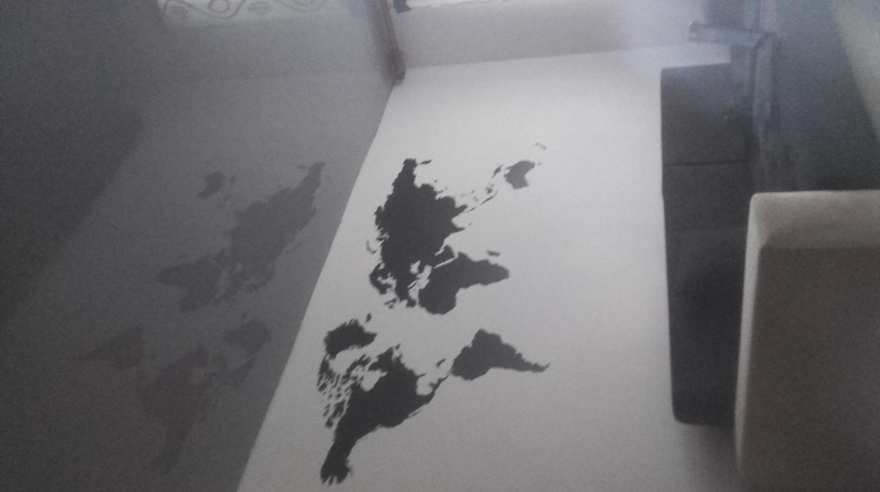 Ściana z namalowaną czarną mapą świata, widoczny fragment narożnika i sofy, słabe oświetlenie.