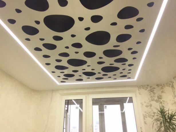 Biały sufit podwieszany z czarnymi, nieregularnymi plamami, obramowany listwą LED, widok na fragment okna i roślinę doniczkową.