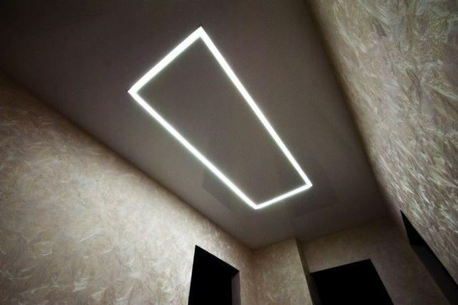 Prostokątna oprawa LED w suficie podwieszanym, ściany z dekoracyjną fakturą, widoczne fragmenty ciemnych otworów drzwiowych.