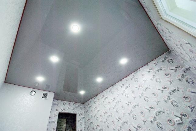 Błyszczący szary sufit napinany z wbudowanymi okrągłymi lampami LED, ściany w jasnej tapecie z motywem podróżniczym, widoczny fragment okna dachowego i kratka wentylacyjna.