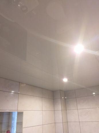 Błyszczący, biały sufit z widocznymi punktowymi lampami w łazience z jasnymi, kwadratowymi płytkami ceramicznymi na ścianach i lustrem.