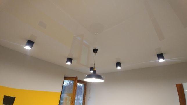 Błyszczący, biały sufit z zainstalowanymi czarnymi, kwadratowymi lampami punktowymi i centralną, wiszącą lampą w stylu industrialnym, widok od dołu.