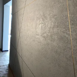 Szara ściana z dekoracyjną, chropowatą fakturą i złotymi liniami, widok z boku, fragment korytarza z białym otworem drzwiowym w tle.