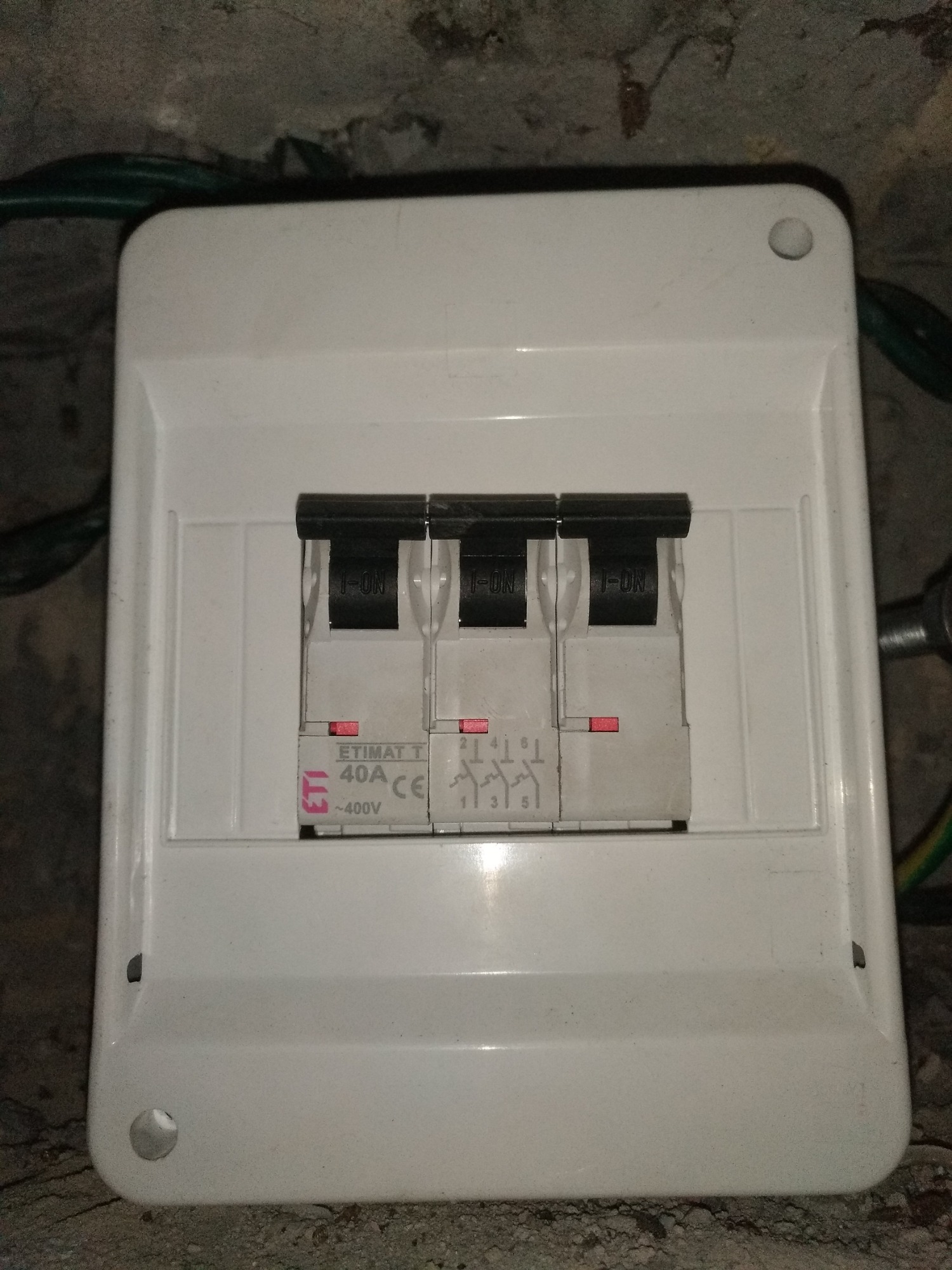 Biała skrzynka z trzema bezpiecznikami ETIMAT 40A w pozycji ON, zamontowana na szarej, surowej ścianie. Widoczne przewody elektryczne w tle.