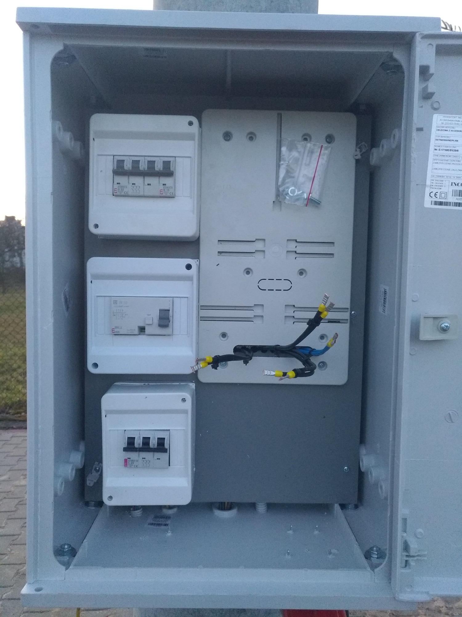 Otwarta skrzynka rozdzielcza z zamontowanymi bezpiecznikami i przewodami z końcówkami kablowymi, widoczne elementy montażowe w opakowaniu foliowym.