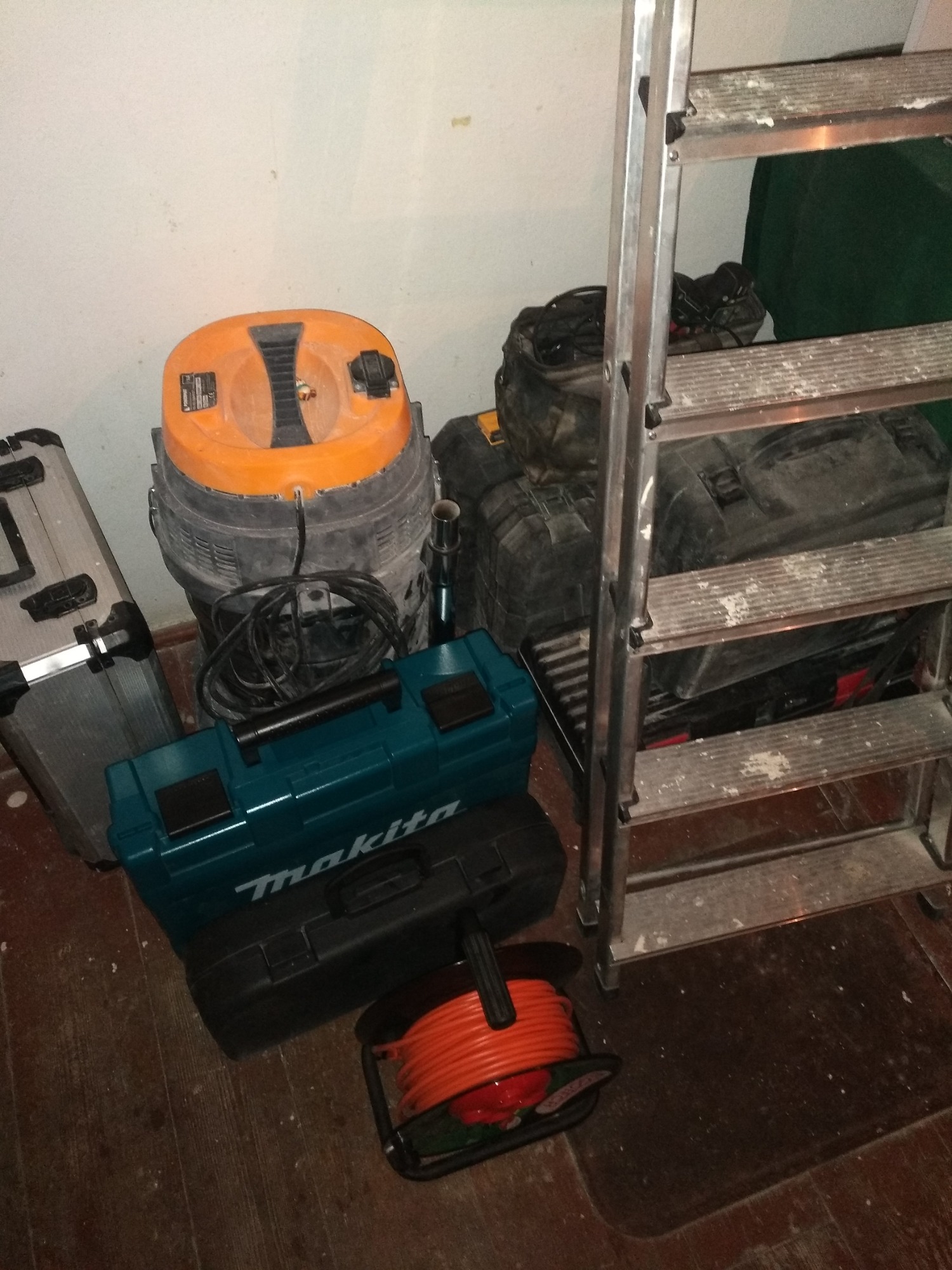 Stojąca aluminiowa drabina, obok niej pomarańczowy odkurzacz przemysłowy, skrzynki narzędziowe Makita i zwinięty pomarańczowy przedłużacz na podłodze w pomieszczeniu.