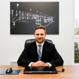 Uśmiechnięty mężczyzna w garniturze siedzi za biurkiem, na którym leży laptop i podkładka pod mysz z napisem 'Business Man', w tle obraz z mostem i okno.