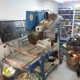 Używana, niebieska piła do cięcia płytek z tarczą diamentową i zanieczyszczonym blatem roboczym, stojąca w warsztacie z regałami narzędziowymi w tle.