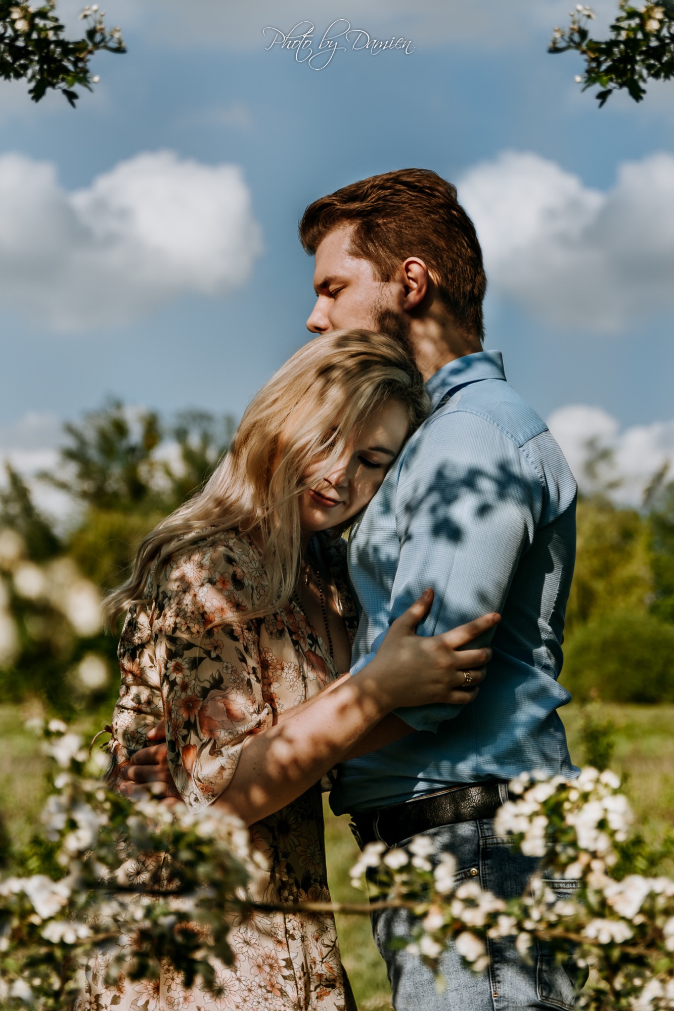 Zakochana para przytulona na tle błękitnego nieba i kwitnących drzew. Kobieta w kwiecistej sukience opiera głowę o ramię mężczyzny w błękitnej koszuli.