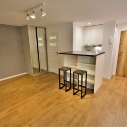 PRO Realizacja - Nowoczesne wnętrze kawalerki po remoncie: szara ściana z płyt, drewniana podłoga, aneks kuchenny z barkiem i stołkami barowymi, przesuwne lustrzane drzwi do szafy.