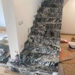 PRO Realizacja - Schody wewnętrzne wyłożone marmurem o wzorze przypominającym fale, widoczne ślady prac wykończeniowych, silikon i puszka farby w tle.