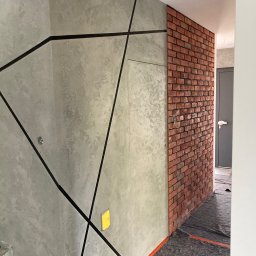 PRO Realizacja - Ściana z dekoracyjną, szarą strukturą i czarnymi liniami tworzącymi geometryczny wzór, sąsiadująca z fragmentem ściany wyłożonej czerwoną cegłą, zabezpieczona podłoga folią ochronną.