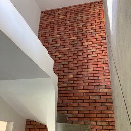 PRO Realizacja - Ściana wykończona czerwoną cegłą klinkierową, widok ze schodów w surowym stanie, z elementami białej konstrukcji stropu i ściany w tle.