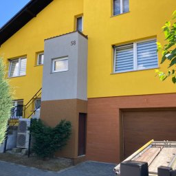 PRO Realizacja - Żółty dom dwukondygnacyjny z szarym pionowym elementem z numerem 58, brązową dolną częścią i garażem, widoczne rusztowanie i zieleń.