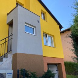 PRO Realizacja - Dwukondygnacyjny dom z żółtą elewacją i szarym gankiem z numerem 58, widoczny klimatyzator Panasonic, brązowe akcenty elewacyjne i zieleń w tle.