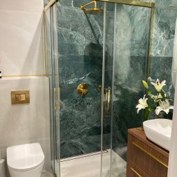 PRO Realizacja - Nowoczesna łazienka z kabiną prysznicową w złotej ramie, marmurowymi ścianami w odcieniach zieleni, wiszącą toaletą i umywalką nablatową z kwiatami.