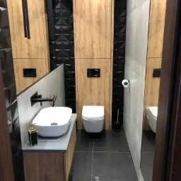 Nowoczesna łazienka w Bielawie z drewnopodobnymi panelami, czarnymi trójwymiarowymi płytkami, wiszącą toaletą i umywalką nablatową, oświetlona wiszącymi lampami.