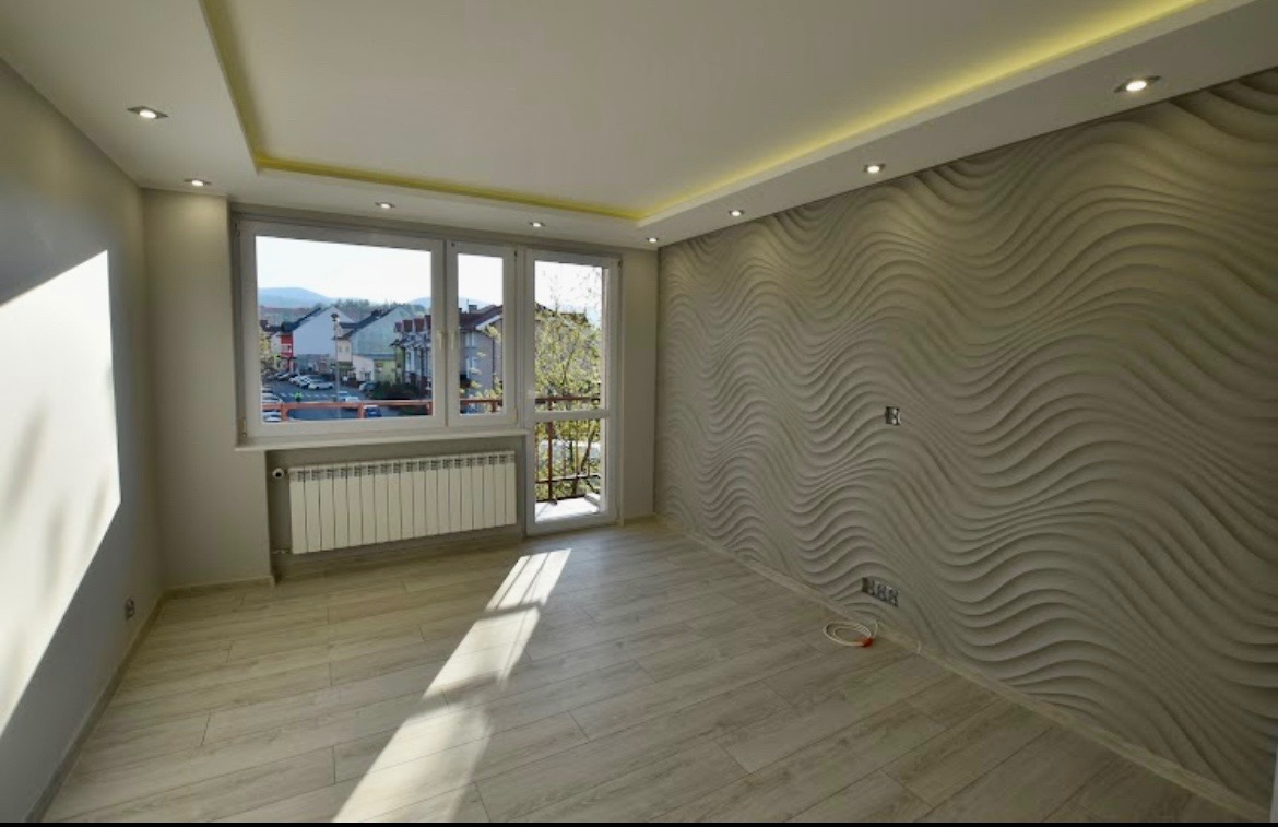 Pusty pokój po remoncie z dekoracyjną ścianą 3D w odcieniach szarości, drewnopodobna podłoga, grzejnik pod oknem i widok na miasto z balkonu.