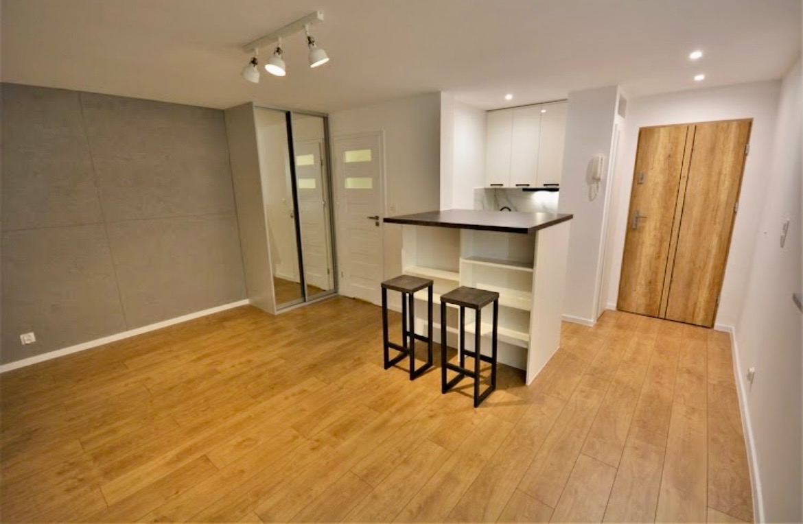 Nowoczesne wnętrze kawalerki po remoncie: szara ściana z płyt, drewniana podłoga, aneks kuchenny z barkiem i stołkami barowymi, przesuwne lustrzane drzwi do szafy.