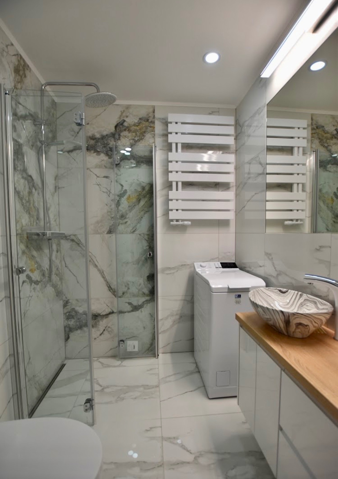 Nowoczesna łazienka z kabiną prysznicową, marmurowymi płytkami, grzejnikiem drabinkowym, pralką i umywalką w geometryczny wzór na drewnianym blacie