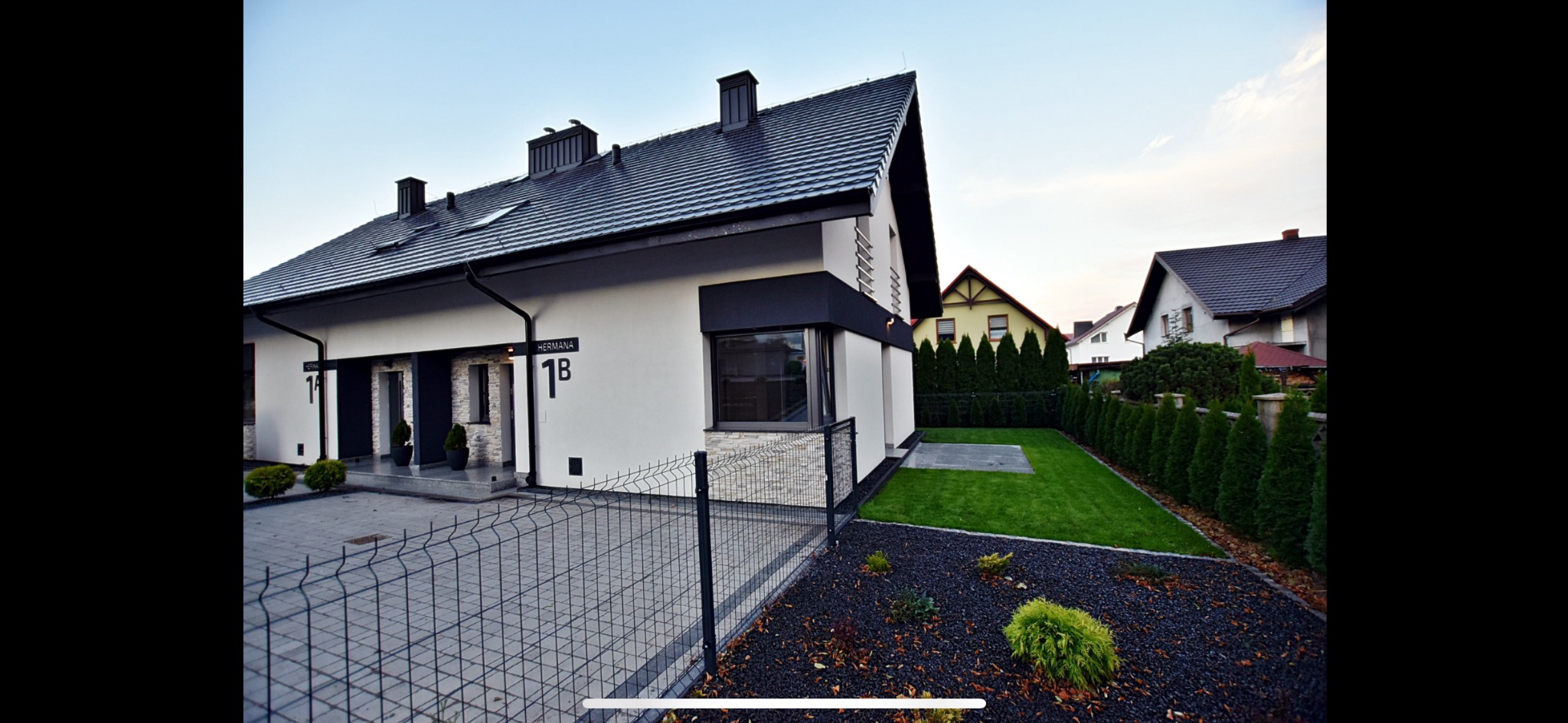 Nowoczesny dom jednorodzinny z białymi ścianami, czarnym dachem i numerem 1B przy wejściu, otoczony zadbanym trawnikiem, ozdobnymi krzewami i czarnym żwirem, widok zza metalowego ogrodzenia.